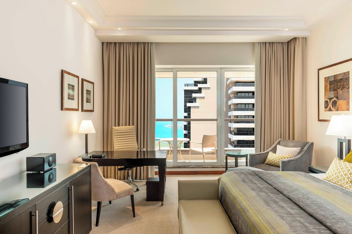 A modern hotel room with a bed, desk, chair, TV, and a balcony with a view of the sea.