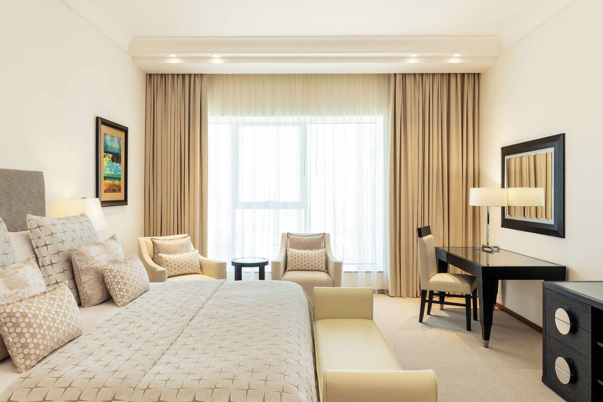 A hotel room with a large bed, two armchairs, a desk, and a large window with curtains.