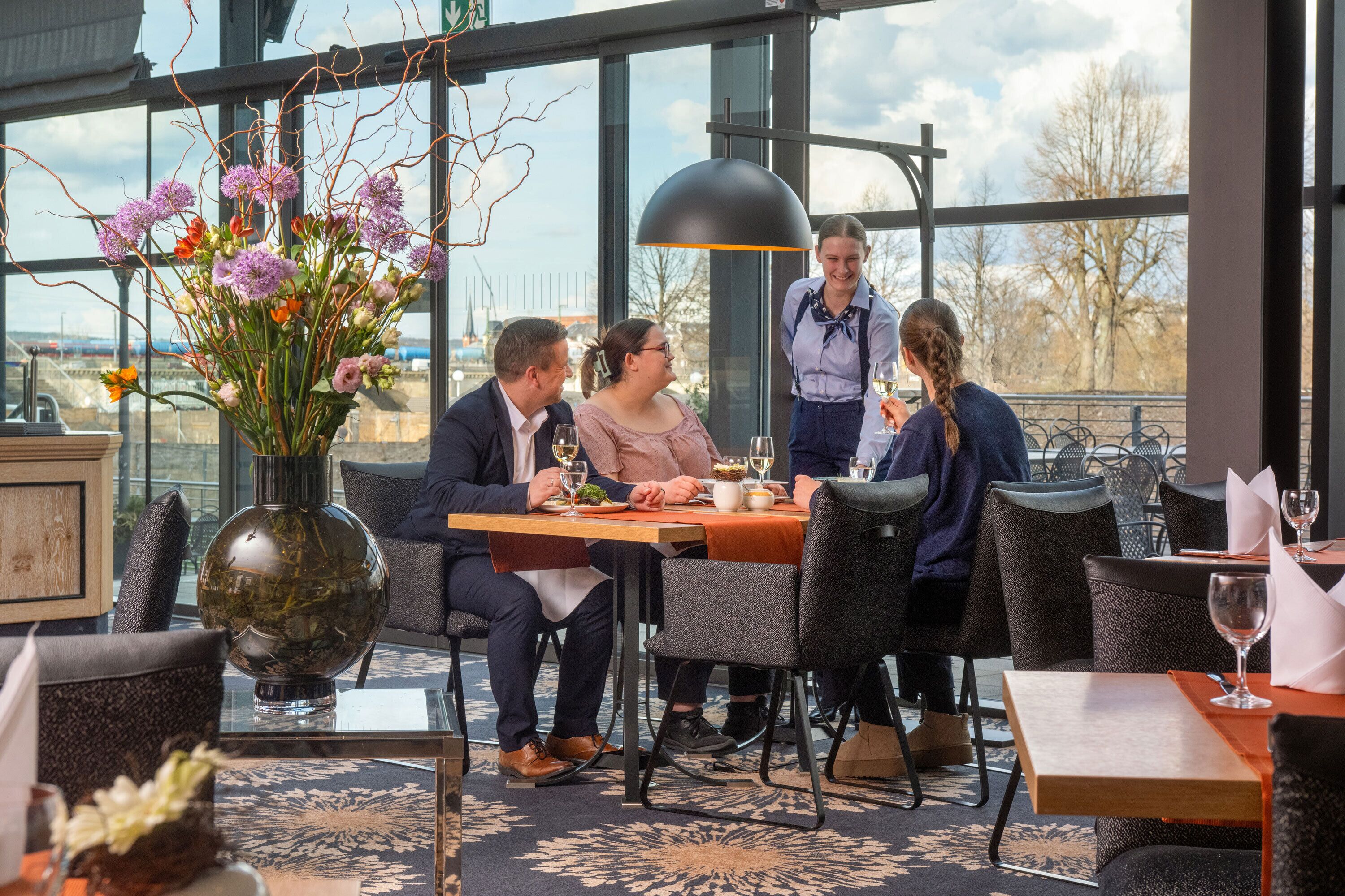 A restaurant interior with a dining table, chairs, a vase of flowers, and a view of the outside.