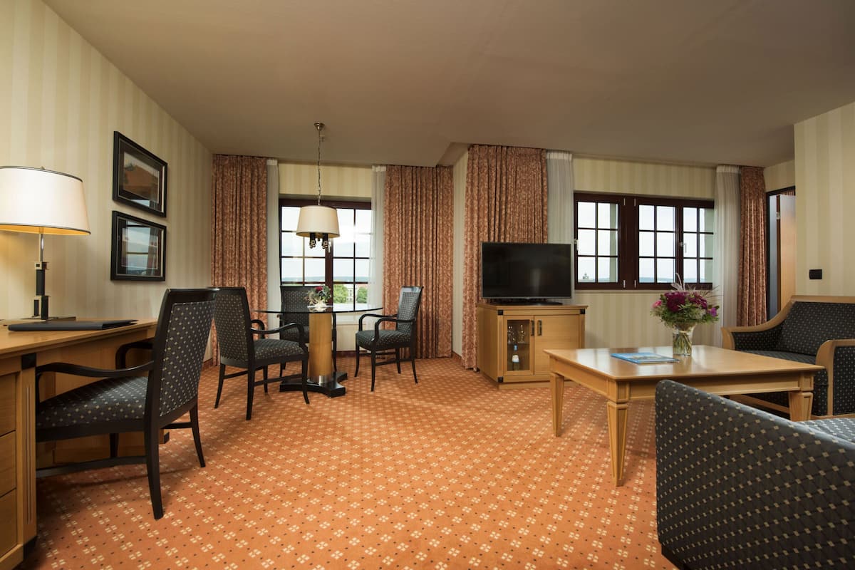 A hotel room with a TV, a desk, two chairs, a sofa, and a small table with a vase of flowers.