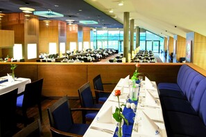 Un amplio comedor con mesas largas, asientos tapizados en azul y grandes ventanales con vistas.