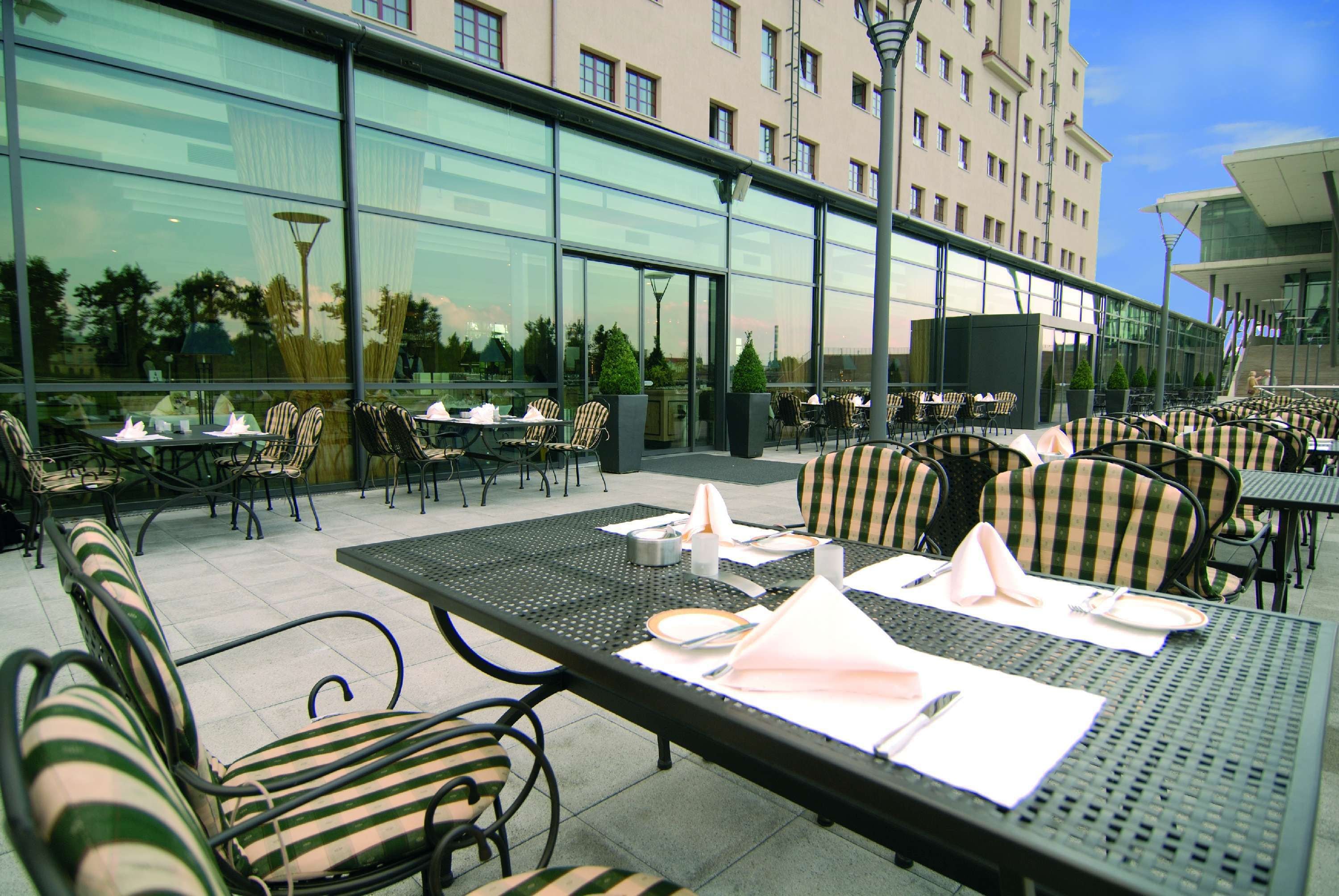 Un área de comedor al aire libre con mesas y sillas, un edificio de cristal al fondo y un cielo despejado.