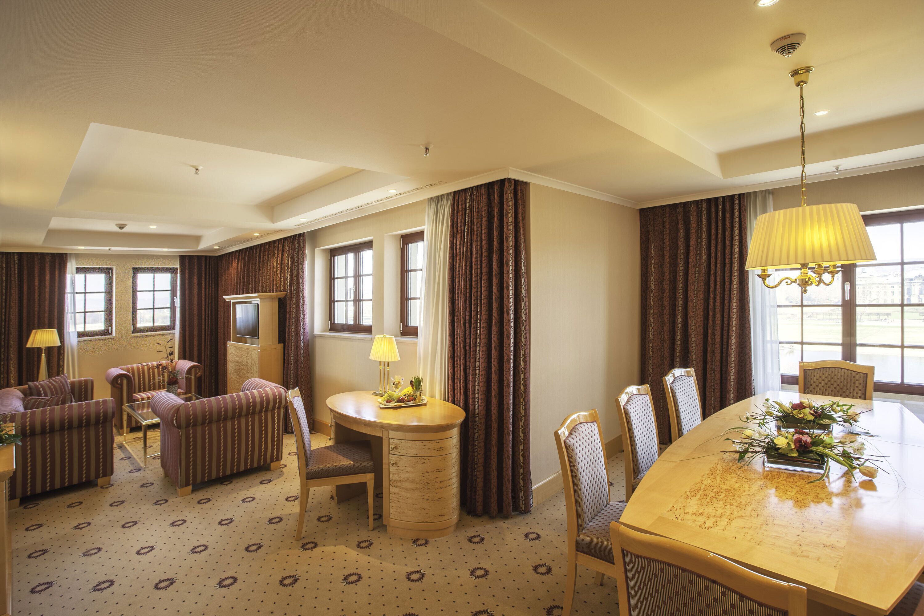 A hotel room with a dining area, a sofa set, a small table with a lamp, and a large window with curtains.