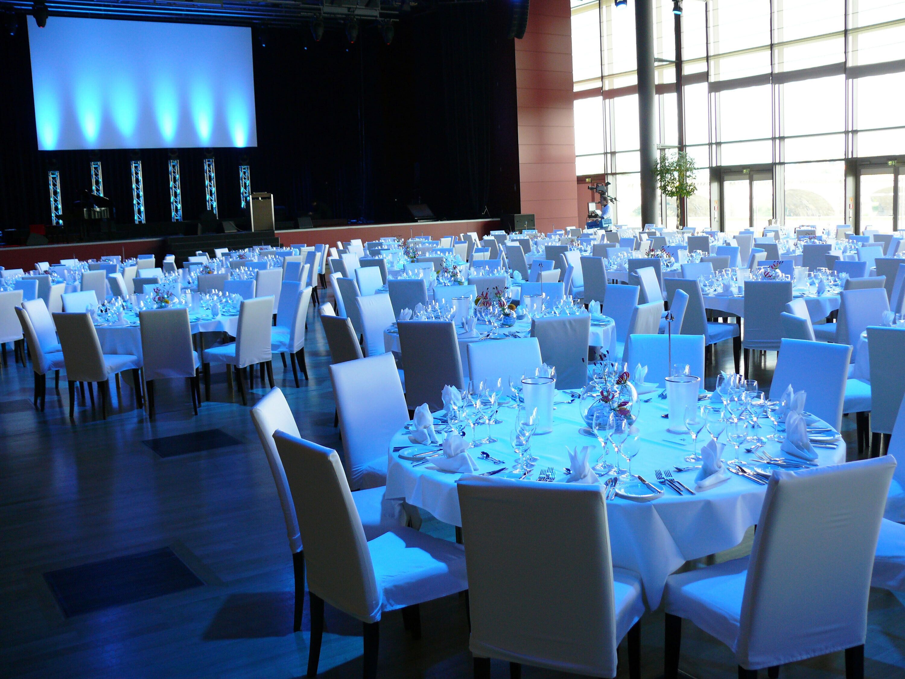 A banquet hall with round tables set for dining, a stage with a screen, and large windows allowing natural light.