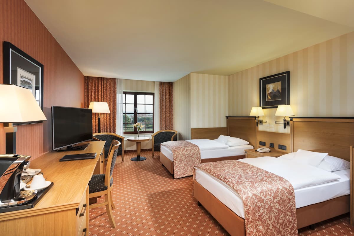 A hotel room with a wooden desk, two beds, a TV, a small table with a lamp, and a window with curtains.