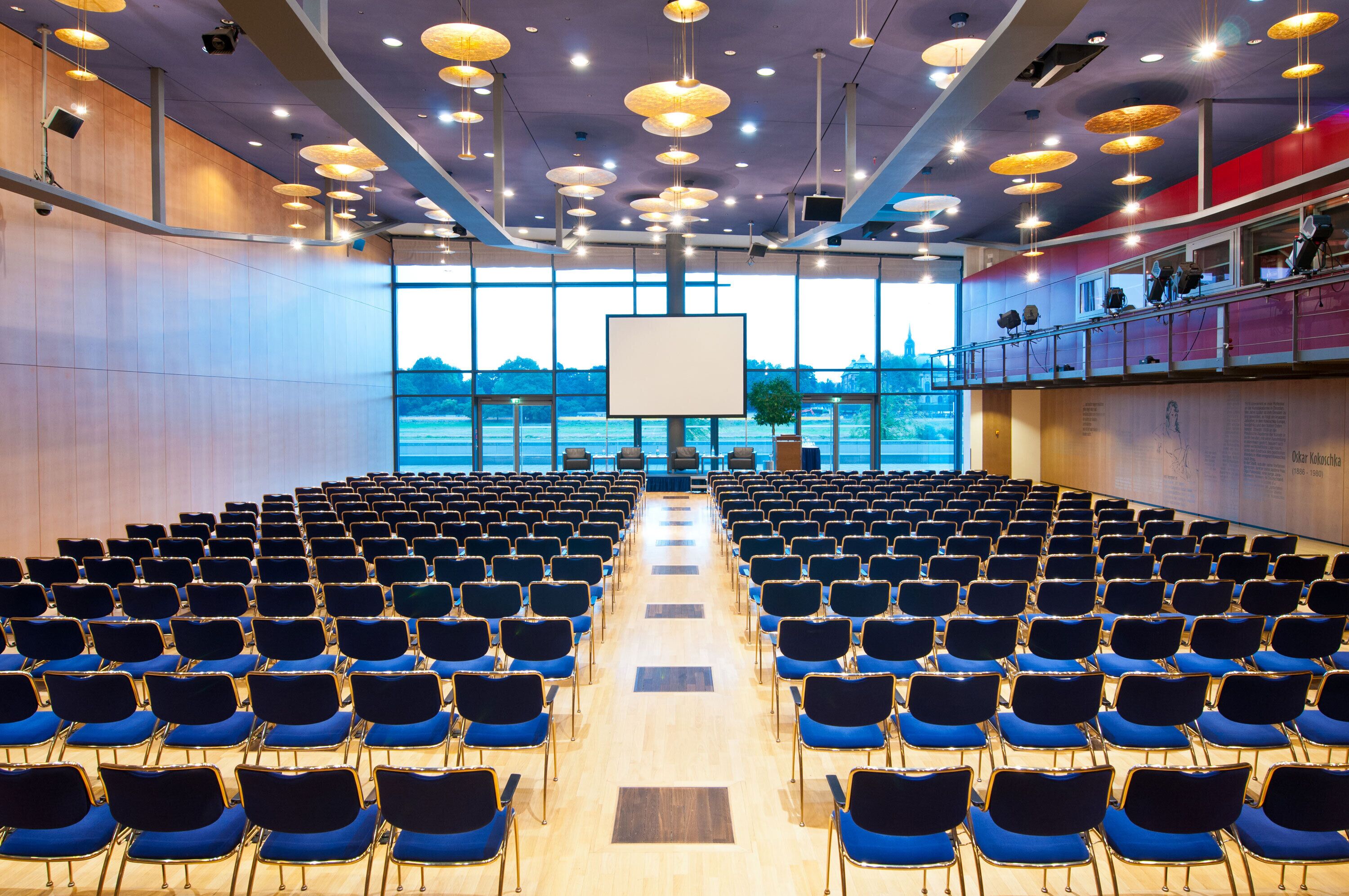 Un salón de conferencias con filas de sillas orientadas hacia una pantalla grande, ventanas amplias y un escenario con equipo de iluminación.