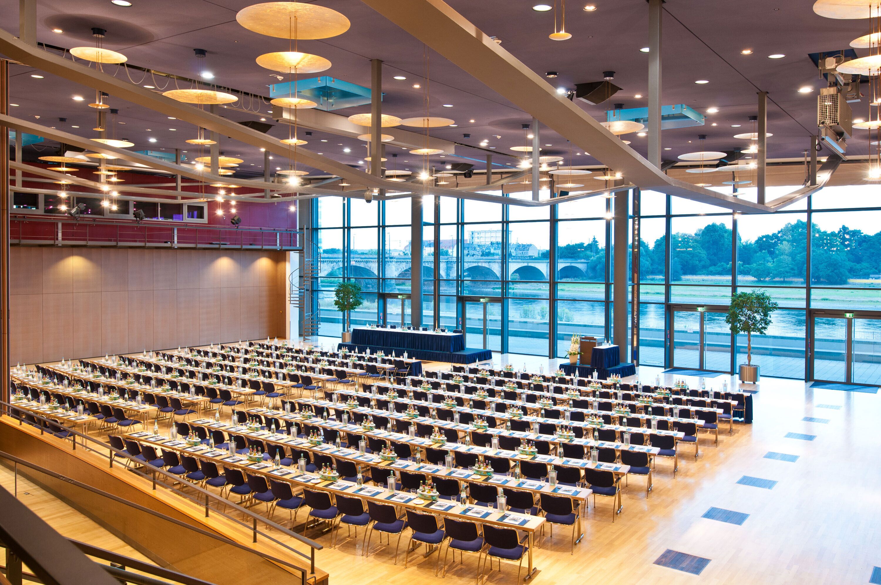 A conference room with rows of tables and chairs, large windows with a view of a bridge and greenery, and modern pendant lighting.