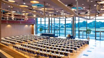 A conference room with rows of tables and chairs, large windows with a view of a bridge and greenery, and modern pendant lighting.