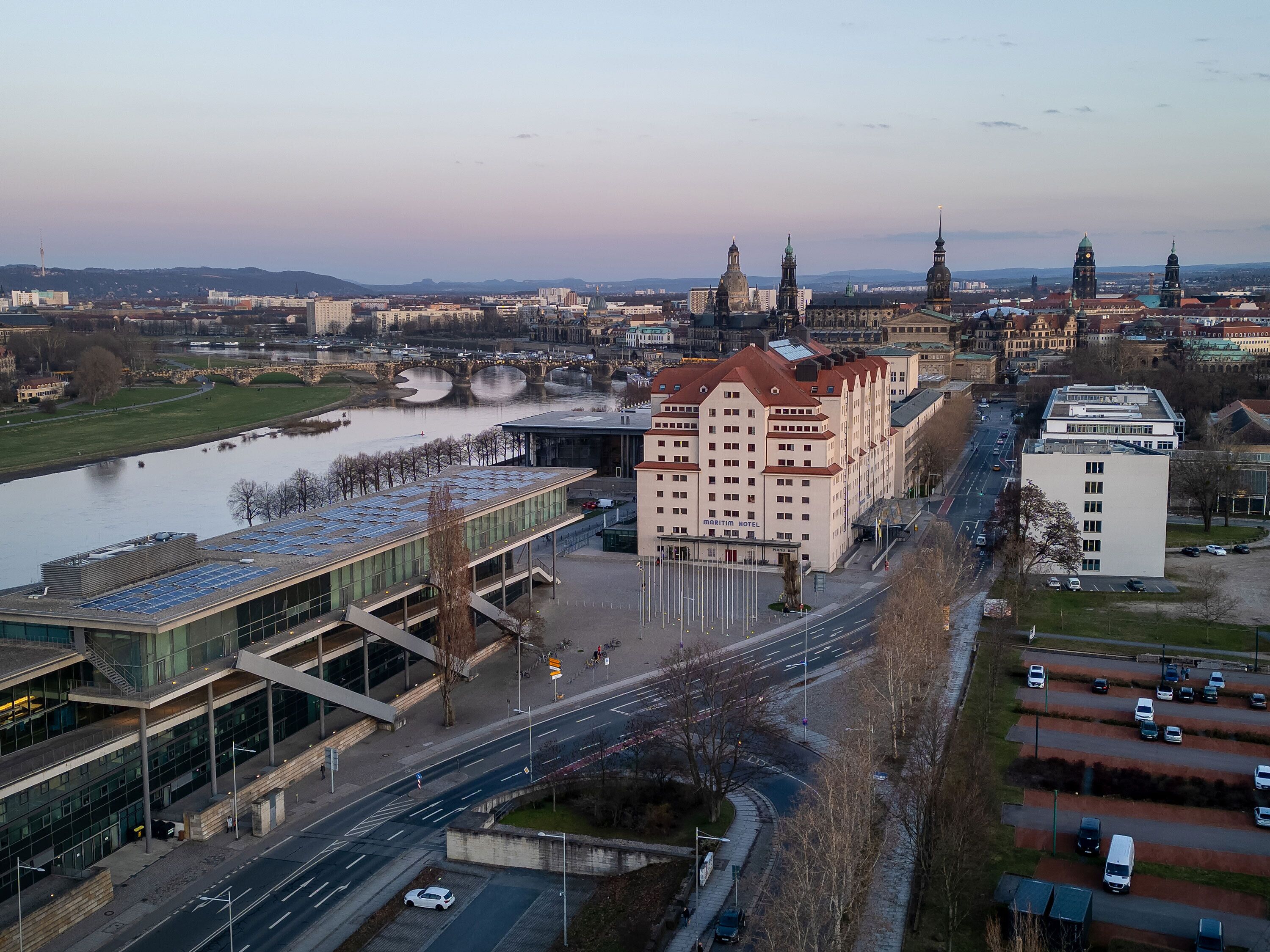 A modern building with a glass facade and a solar panel roof, adjacent to a river and a historic building with a red roof.