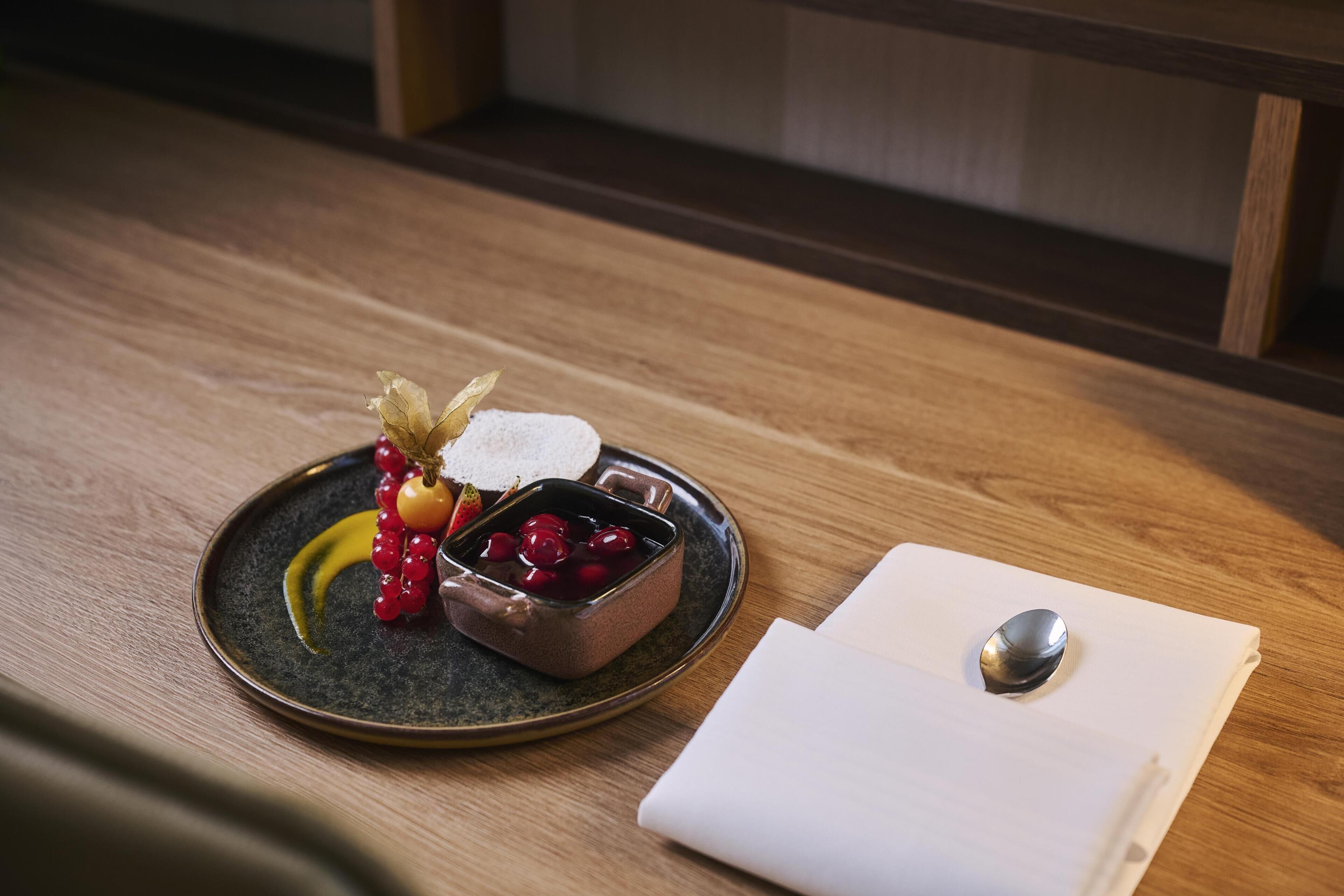 A wooden table with a ceramic plate containing red currants and a small dish of jam, a spoon, and a napkin.