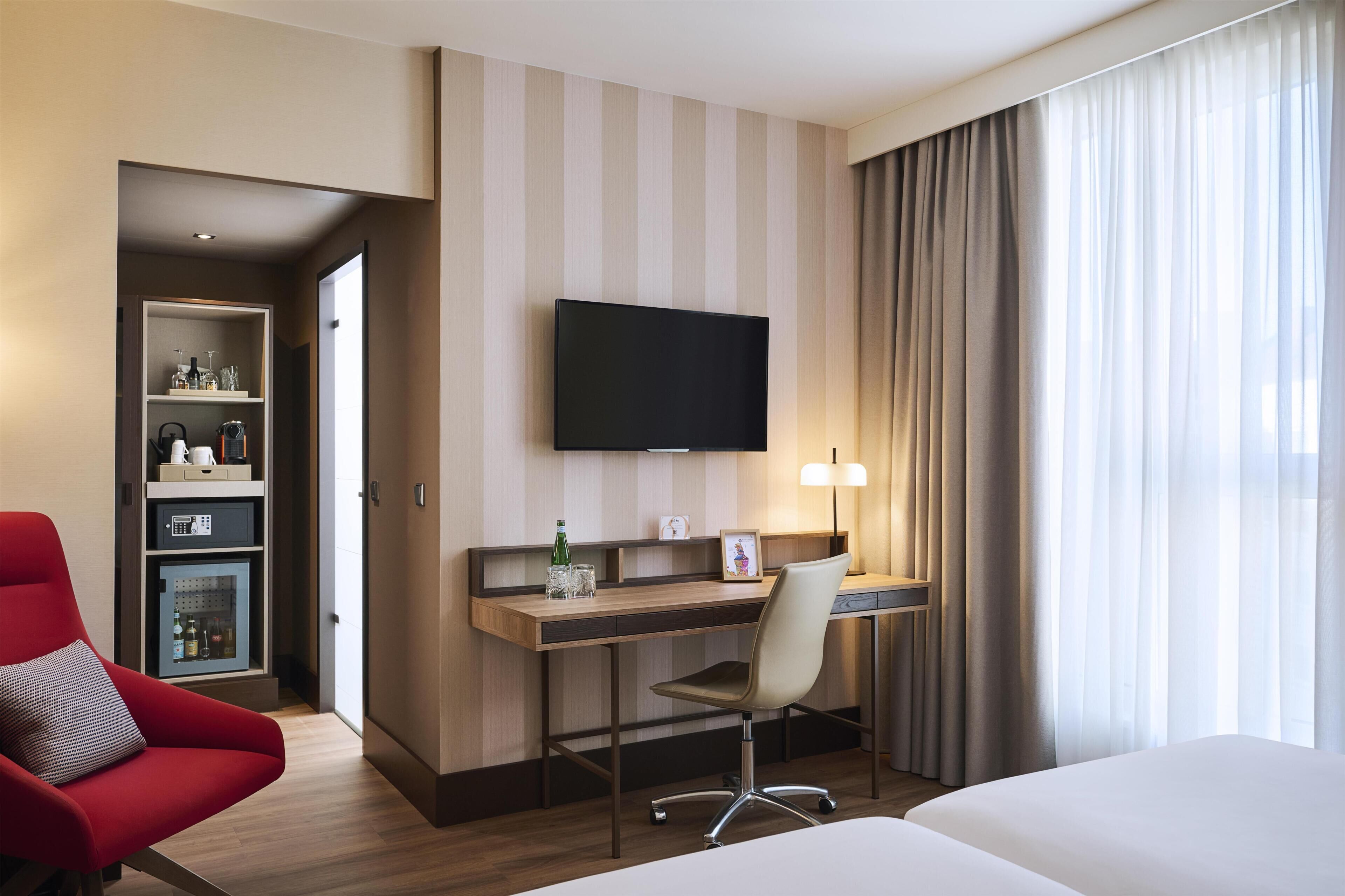 Habitación de hotel con cama, escritorio con silla, televisor, lámpara y minibar.