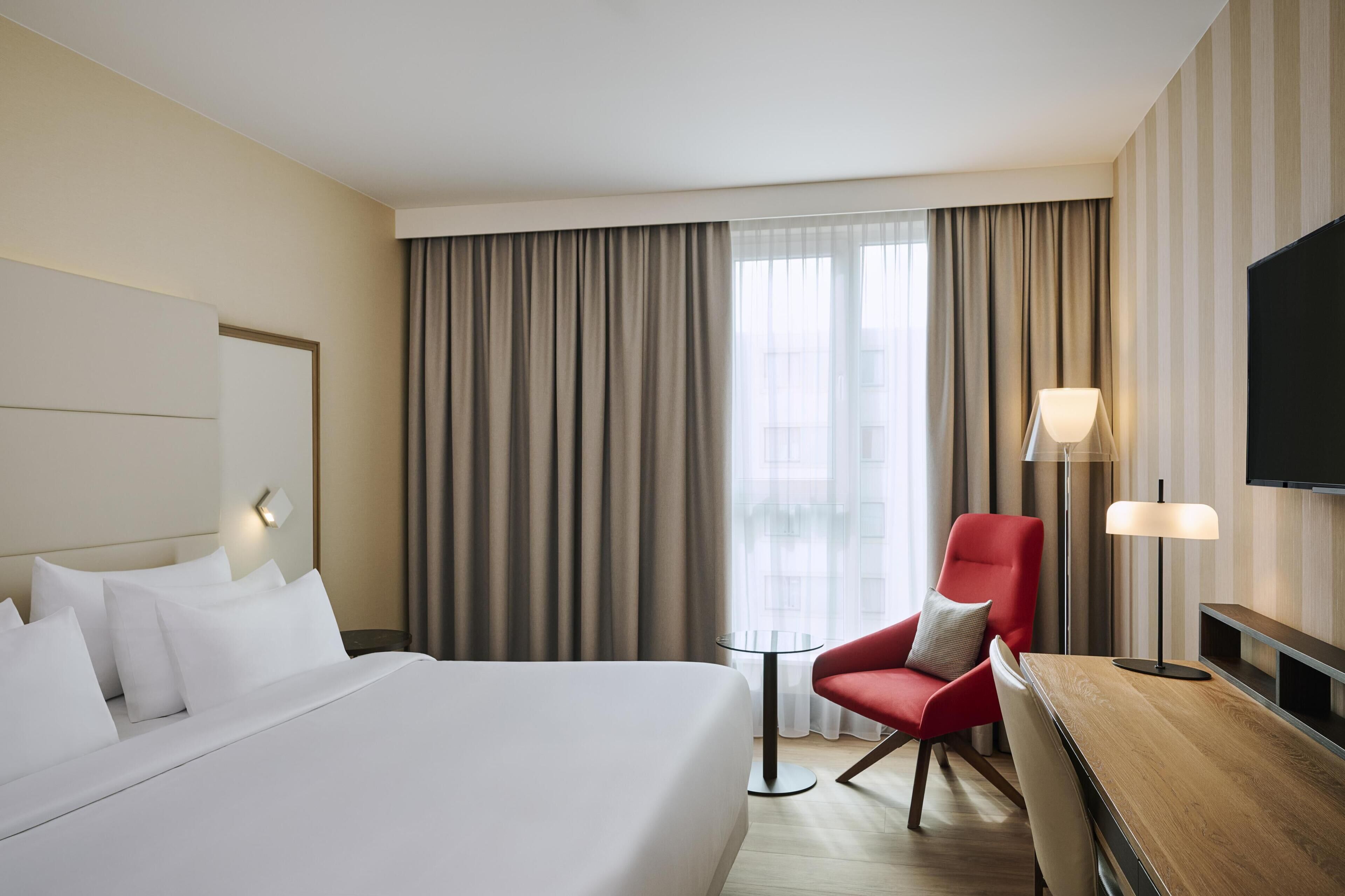 Habitación de hotel con una cama grande, un sillón rojo, una mesita, un televisor y un ventanal con cortinas.