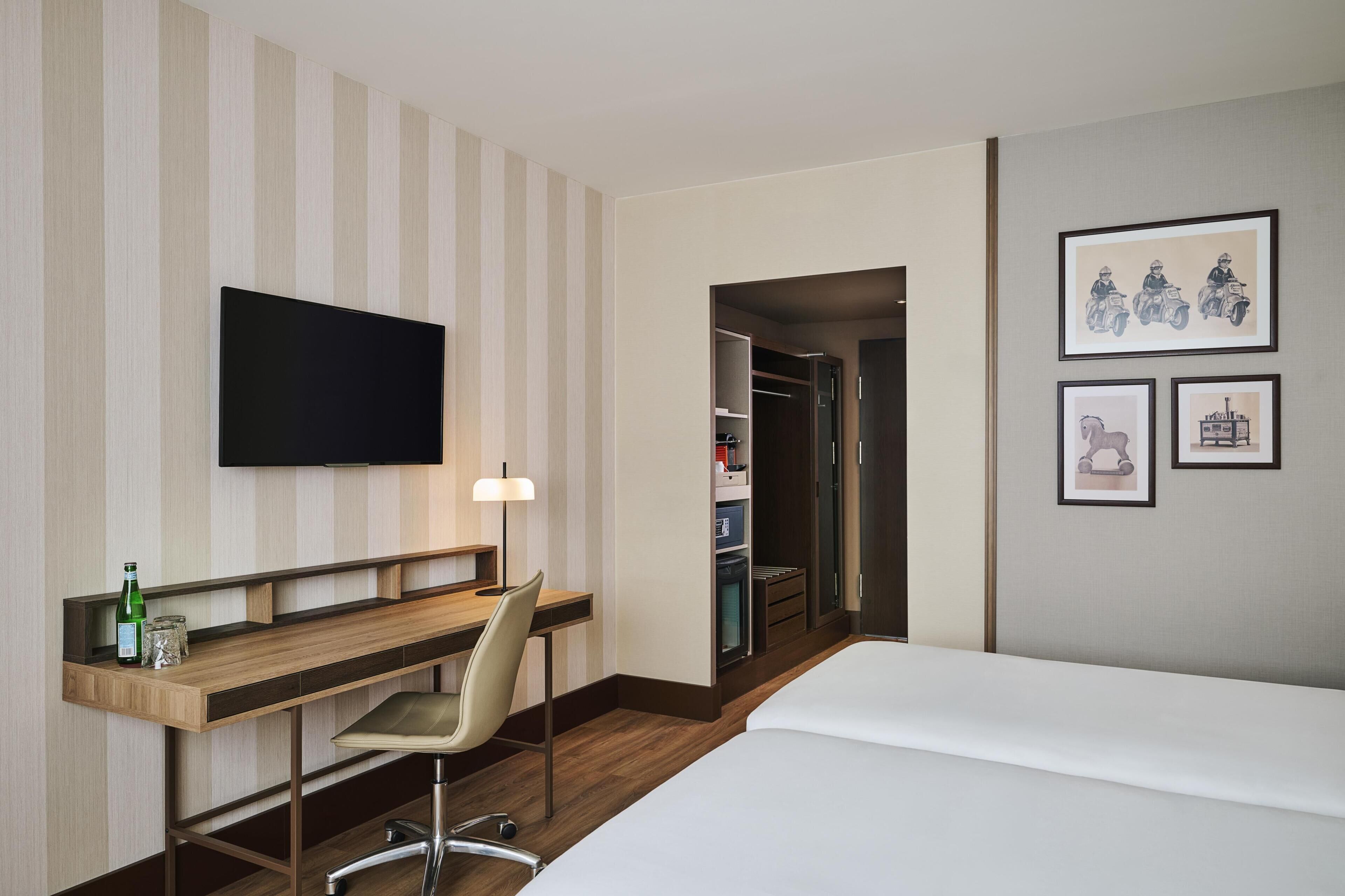 A hotel room with a bed, a desk with a chair, a television, and framed pictures on the wall.