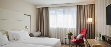 A hotel room with two beds, a red chair, a small table with a lamp, and a TV on the wall.