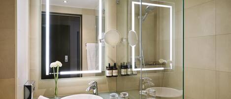 Un baño moderno con tocador de vidrio, un espejo grande, cabina de ducha con vidrio y lavamanos blanco.
