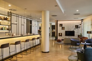 A modern bar area with a long counter, multiple stools, and a well-stocked bar. There is a seating area with sofas and chairs, and a television mounted on the wall.