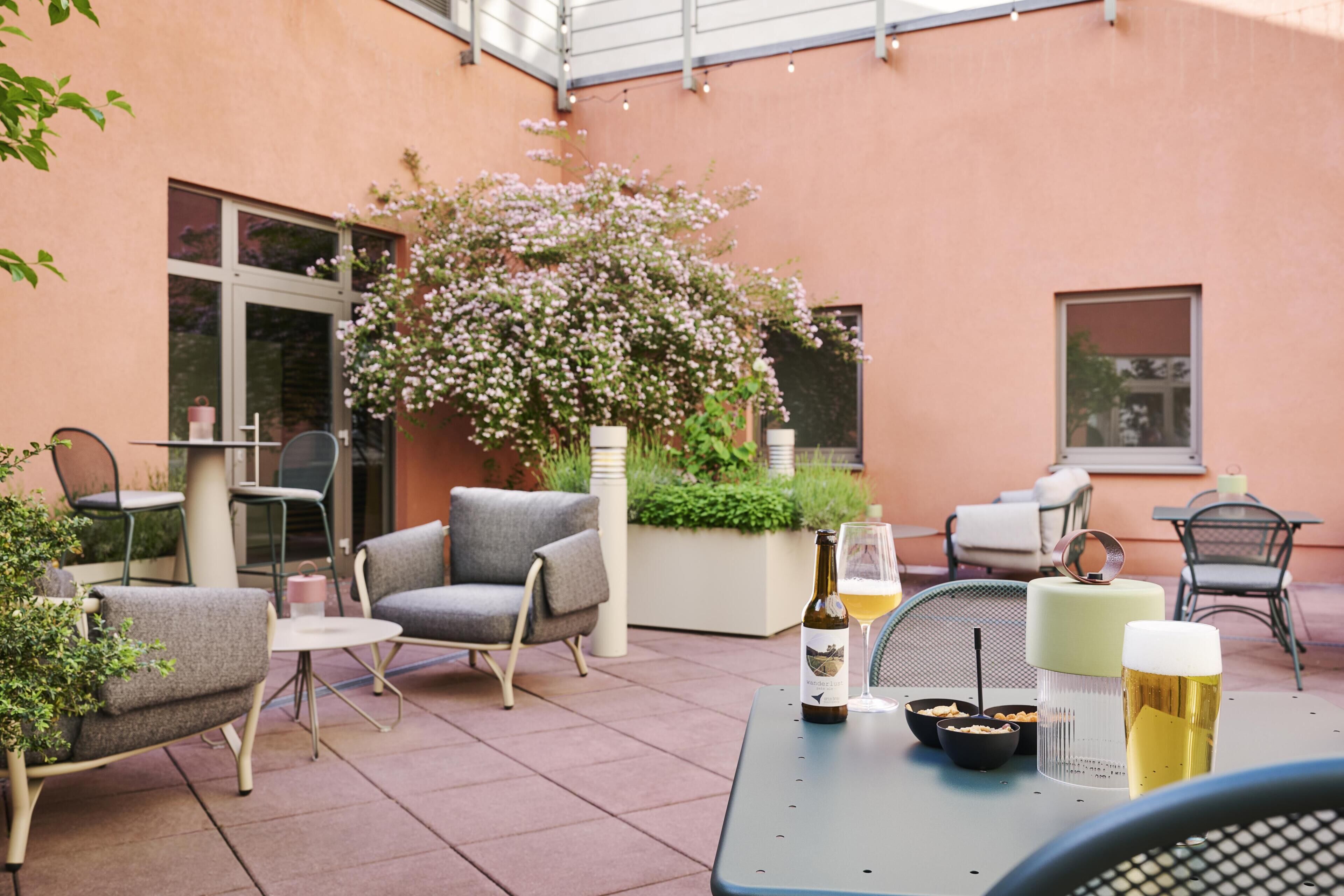 Een terras buiten met zitplaatsen, een tafel met een fles en glazen, en een bloemenstruik.