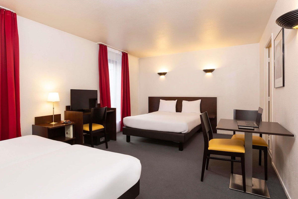 A hotel room with two beds, a desk with a laptop, a chair, a lamp, and two wall-mounted lights.