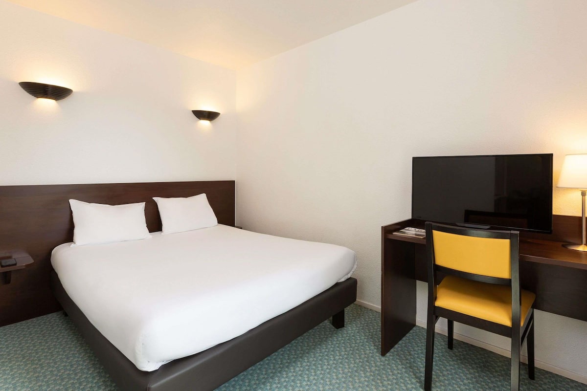 A hotel room with a bed, a desk, a chair, a television, and two wall-mounted lights.