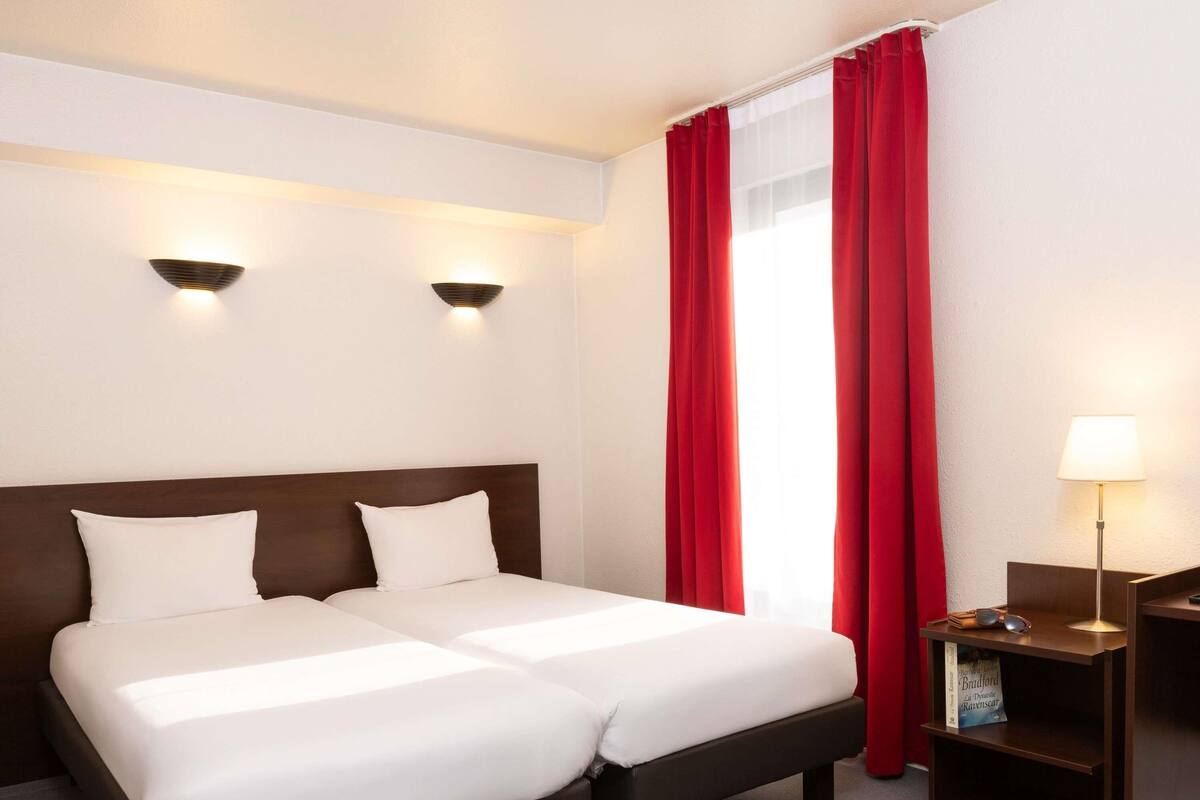 Quarto de hotel com cama de casal, duas luminárias fixadas na parede, mesa de cabeceira com abajur e cortina vermelha.