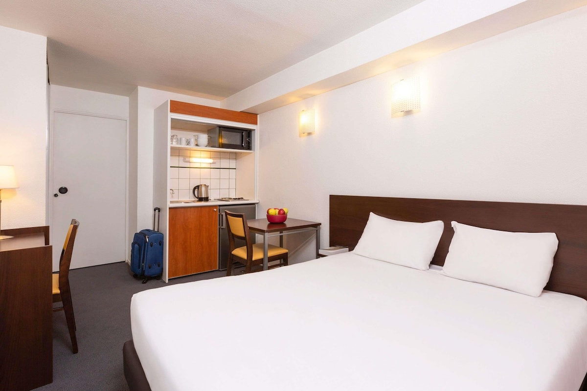 A hotel room with a bed, a desk with a microwave, a chair, and a suitcase.
