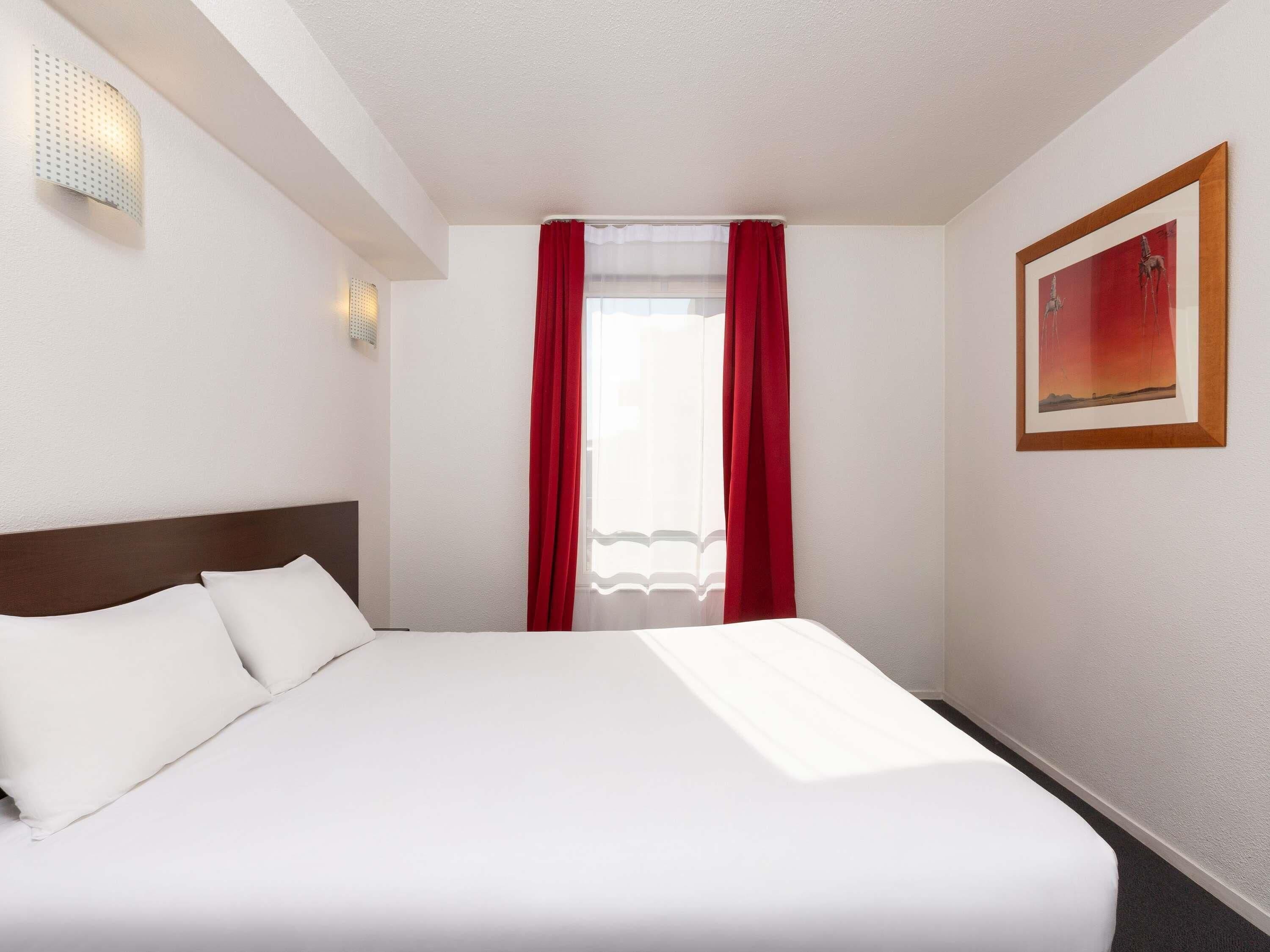 A hotel room with a bed, a wall-mounted light, a window with red curtains, and a framed picture on the wall.