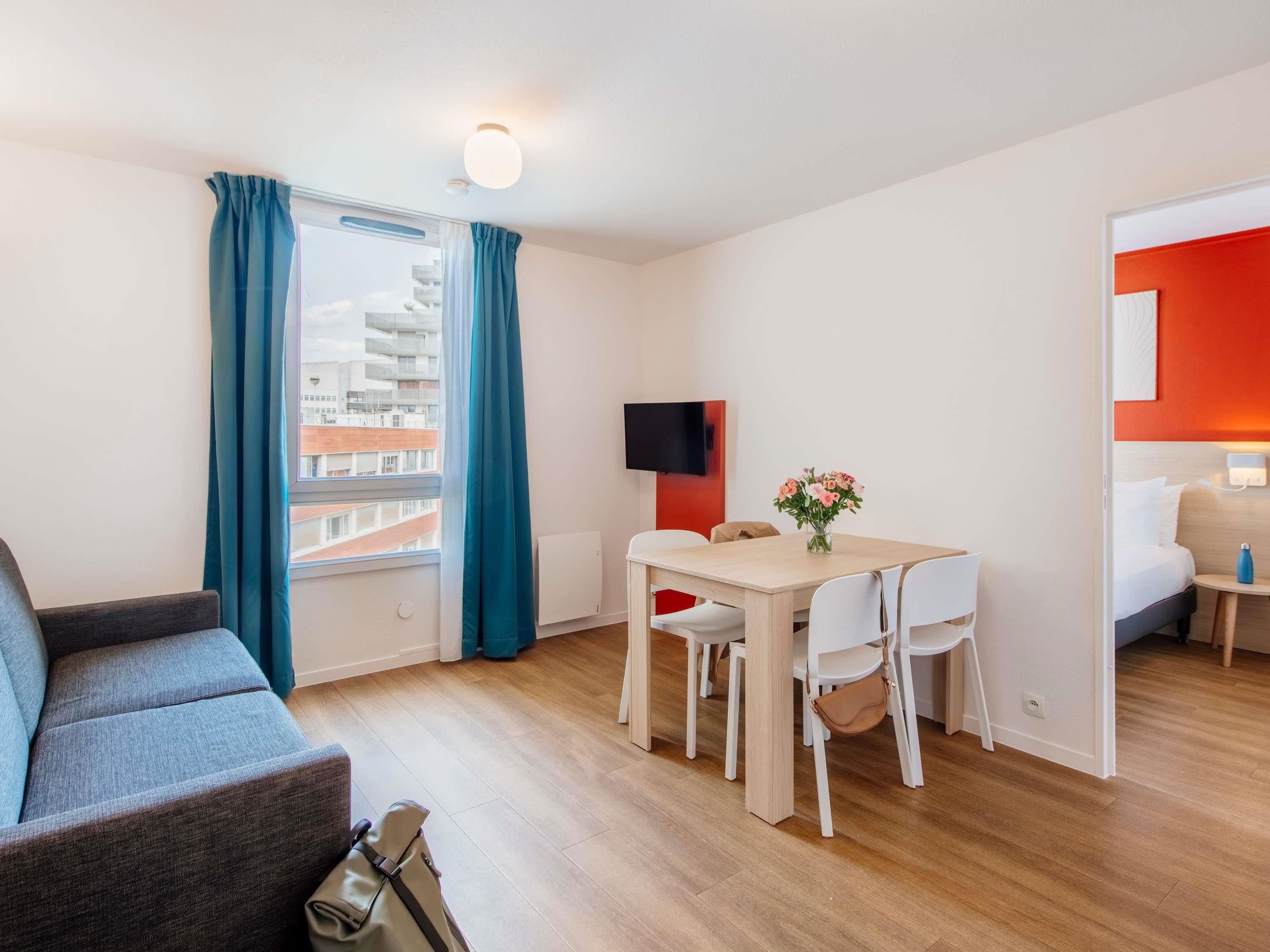 A modern hotel room with a sofa, a dining table with chairs, a TV, and a bed.