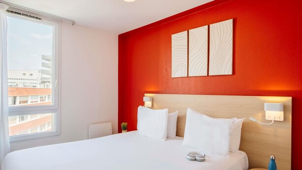 Hotelkamer met een groot bed, een klein nachtkastje, een wandlamp en een raam met uitzicht op gebouwen.