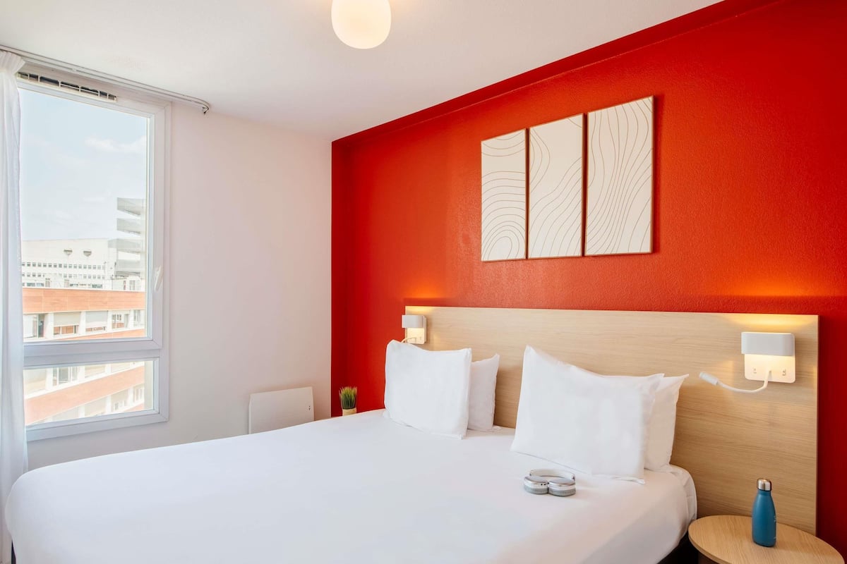 Ein Hotelzimmer mit einem großen Bett, einem kleinen Nachttisch, einer Wandleuchte und einem Fenster mit Blick auf Gebäude.