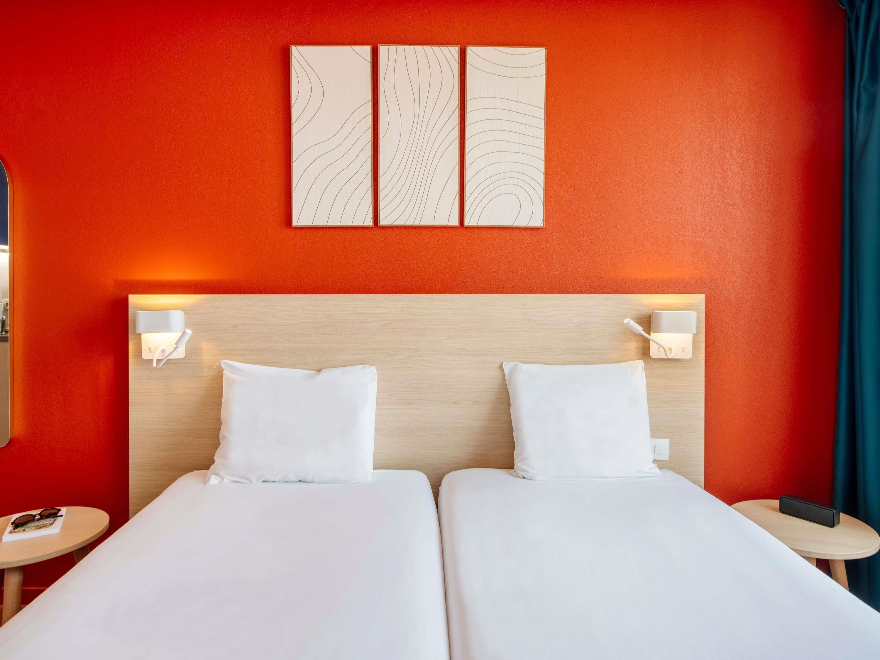 Camera d'albergo con due letti, una testiera in legno e una parete rossa con un pannello decorativo.
