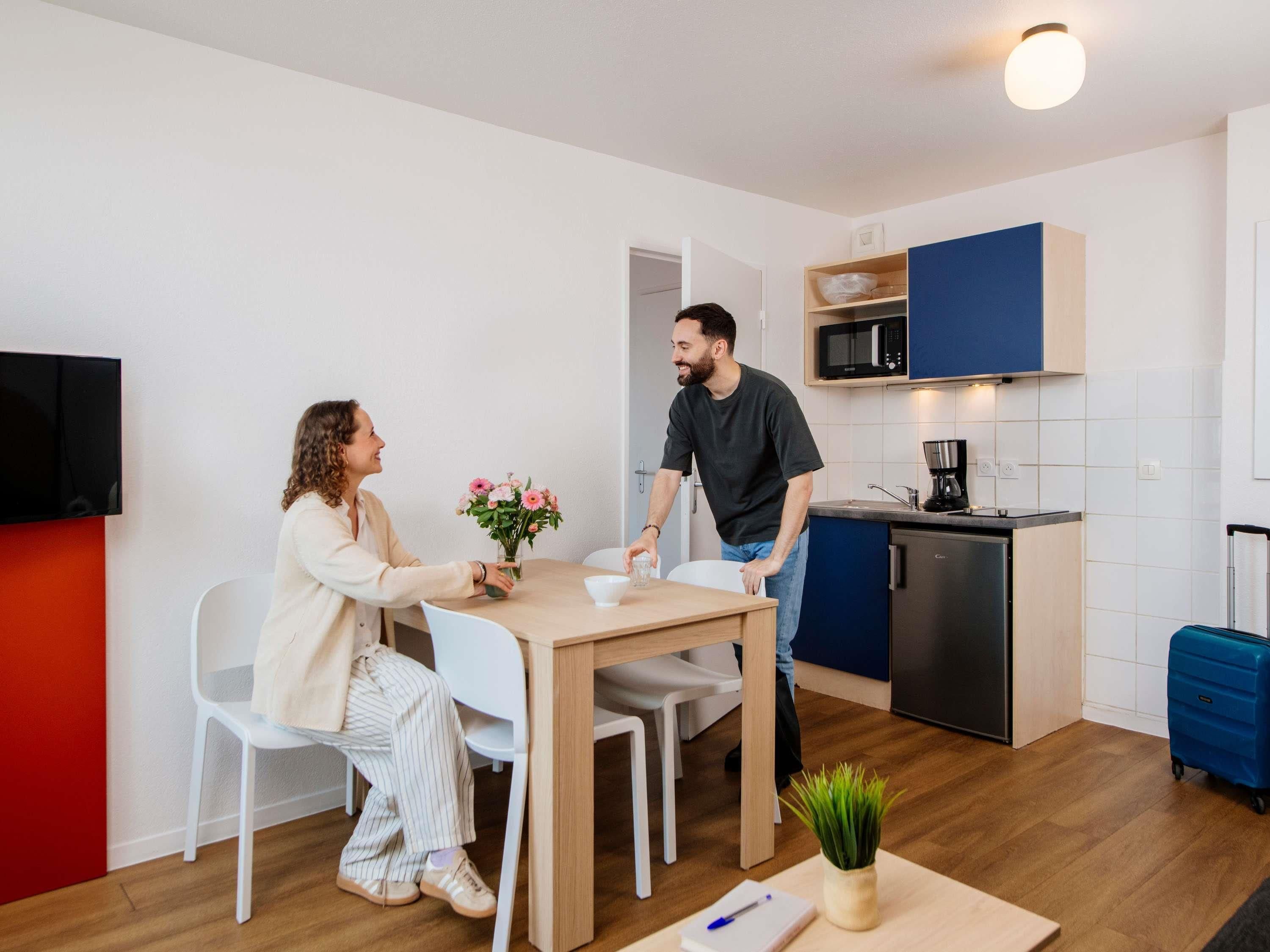 A modern kitchen with a microwave, coffee maker, and a small refrigerator. There is a dining table with chairs, a potted plant, and a suitcase on the floor.