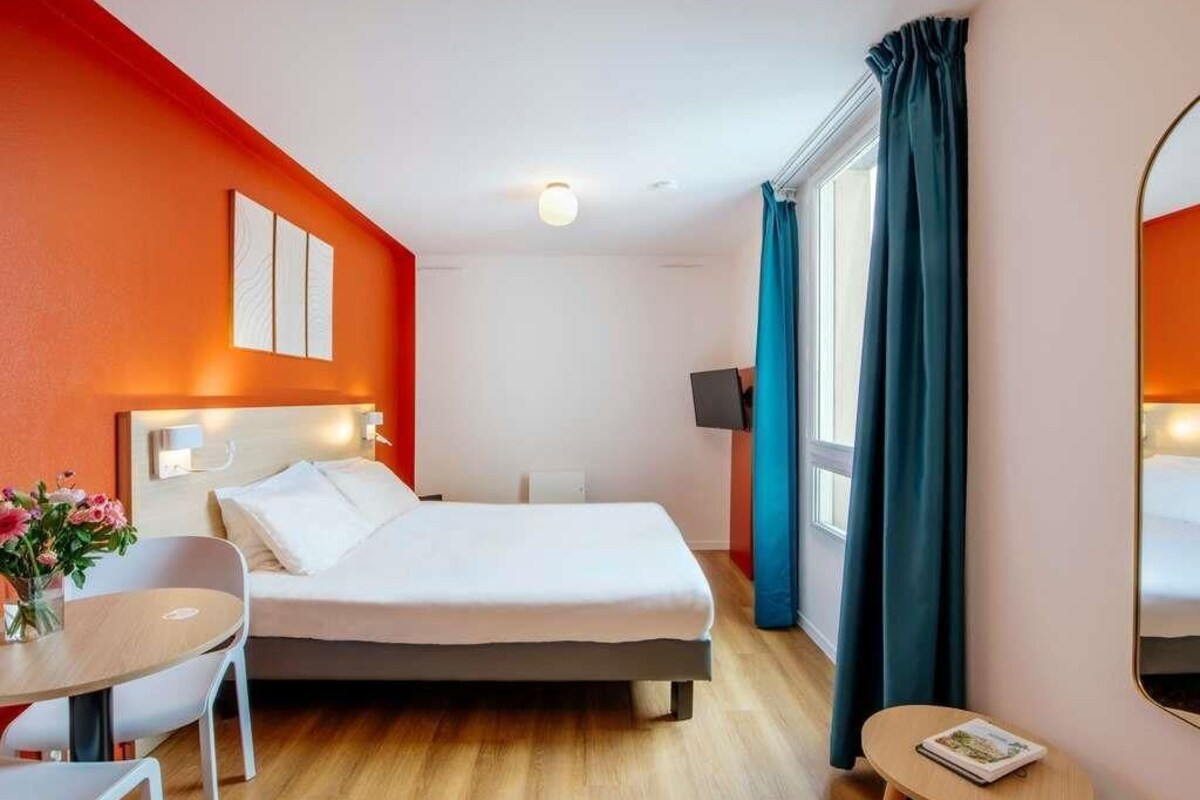 A hotel room with a bed, a desk, a chair, a small table, a mirror, and a window with curtains.