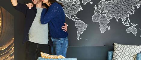 A couple taking a selfie in front of a world map with 'THE ADAGIO COMMUNITY' text. There's a round table with a book and a cup, and a blue chair with patterned cushions.