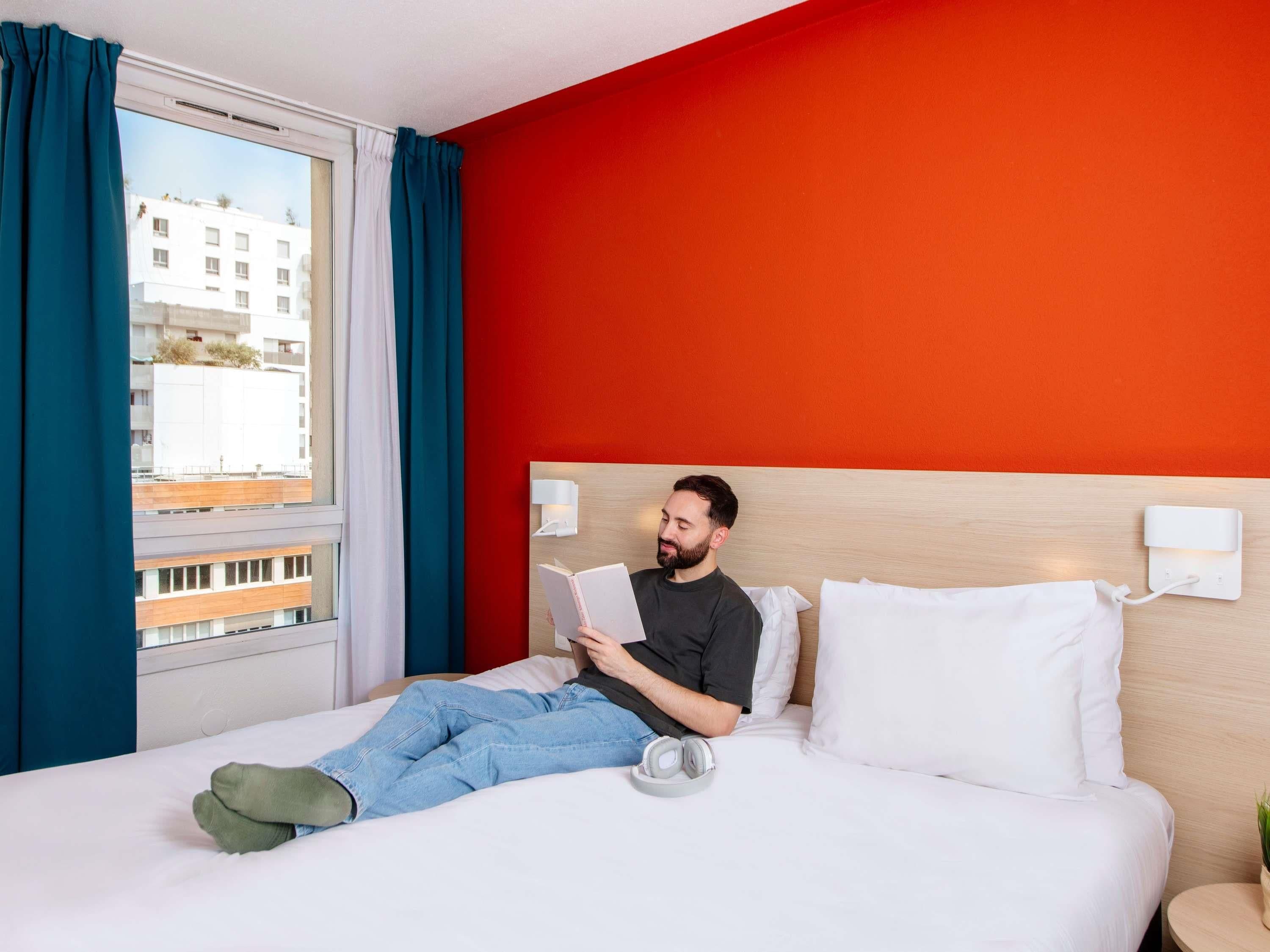 Una camera d'albergo con un letto, una lampada da comodino, una finestra con tende e vista sugli edifici.