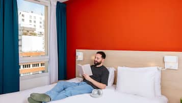 Una camera d'albergo con un letto, una lampada da comodino, una finestra con tende e vista sugli edifici.
