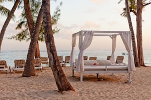 Un paisaje playero con una cama con dosel, sillas de playa y palmeras.