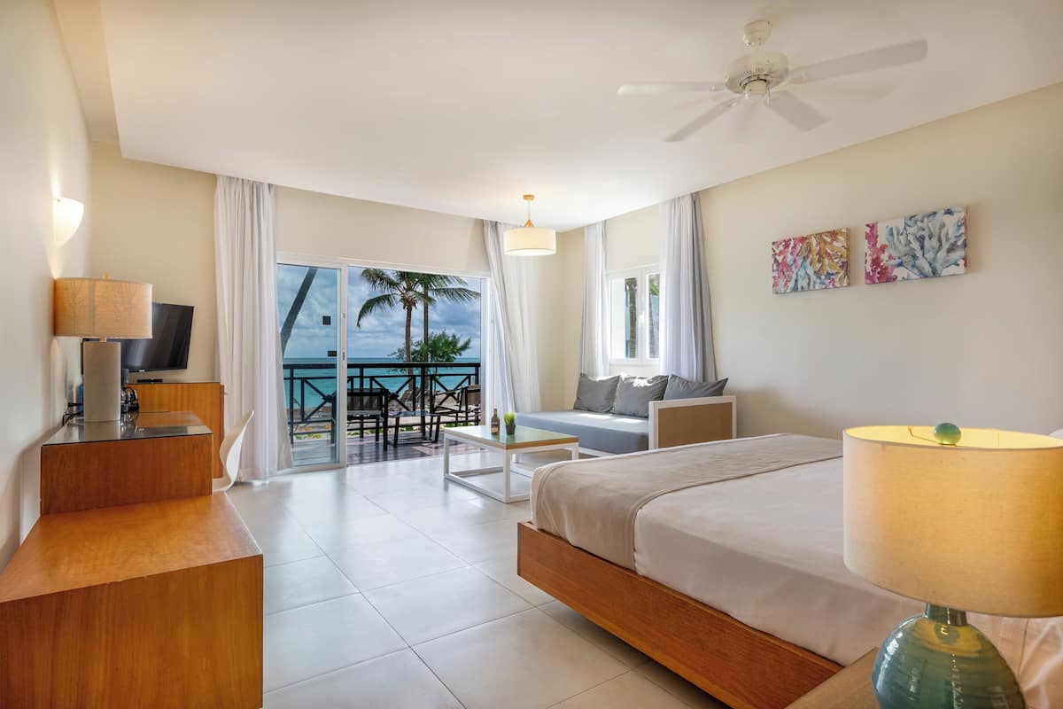 A modern hotel room with a bed, a desk, a chair, a sofa, a balcony with a view, and a ceiling fan.
