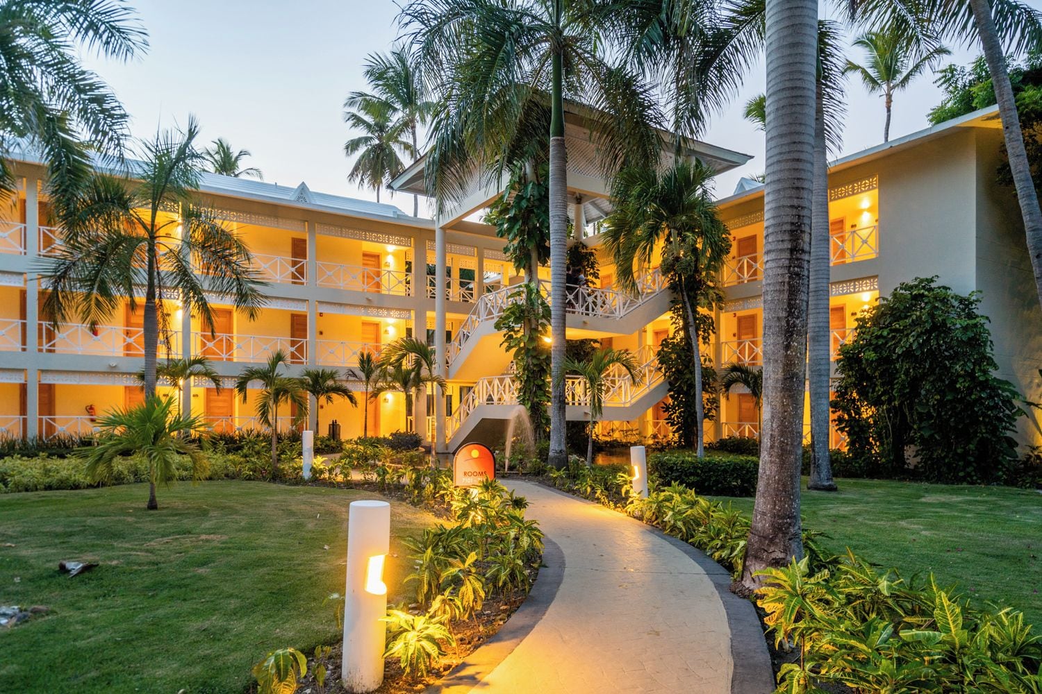 Un hotel de varios pisos con un patio central, palmeras y un camino que lleva a la entrada.