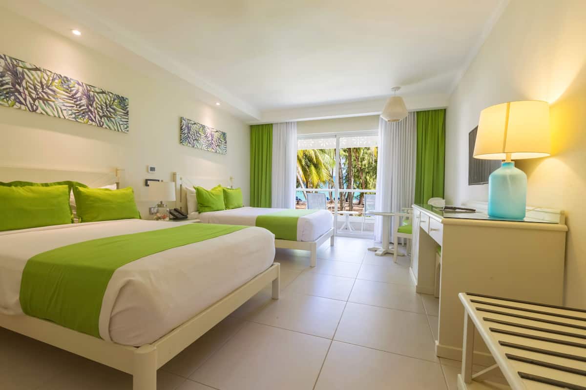 A hotel room with two beds, a desk, a chair, a lamp, and a balcony with a view of palm trees.