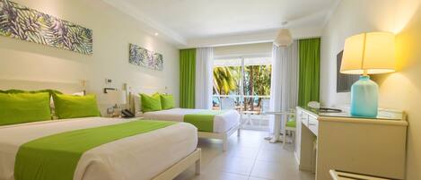 Hotelkamer met twee bedden, een bureau, een stoel, een lamp en een balkon met uitzicht op palmbomen.