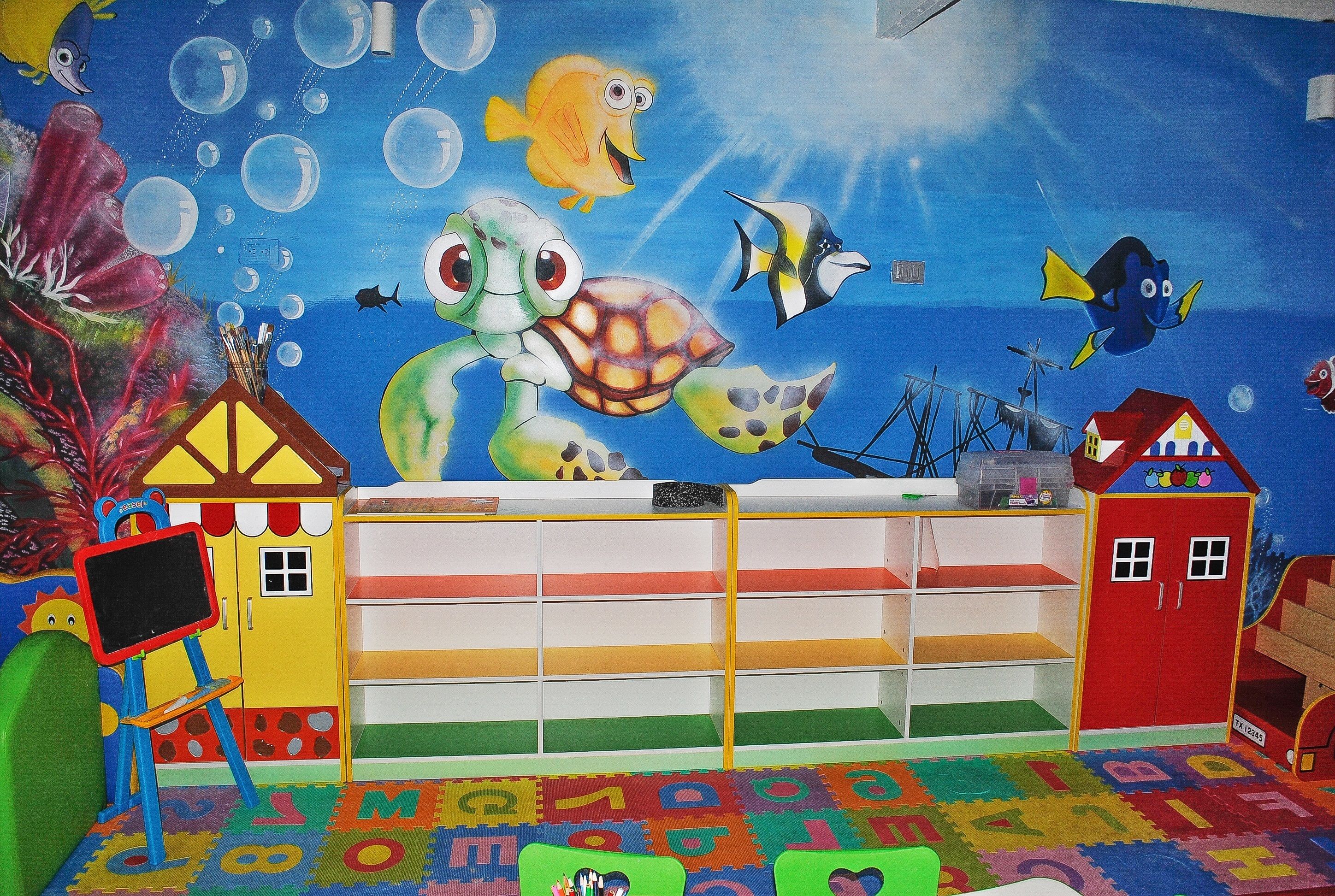 Un salón infantil con un mural colorido, un pequeño escenario y una estantería.