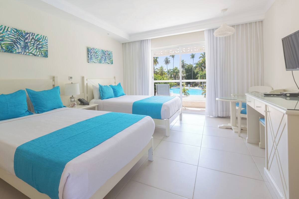 A hotel room with two beds, a desk, and a view of a pool and palm trees.