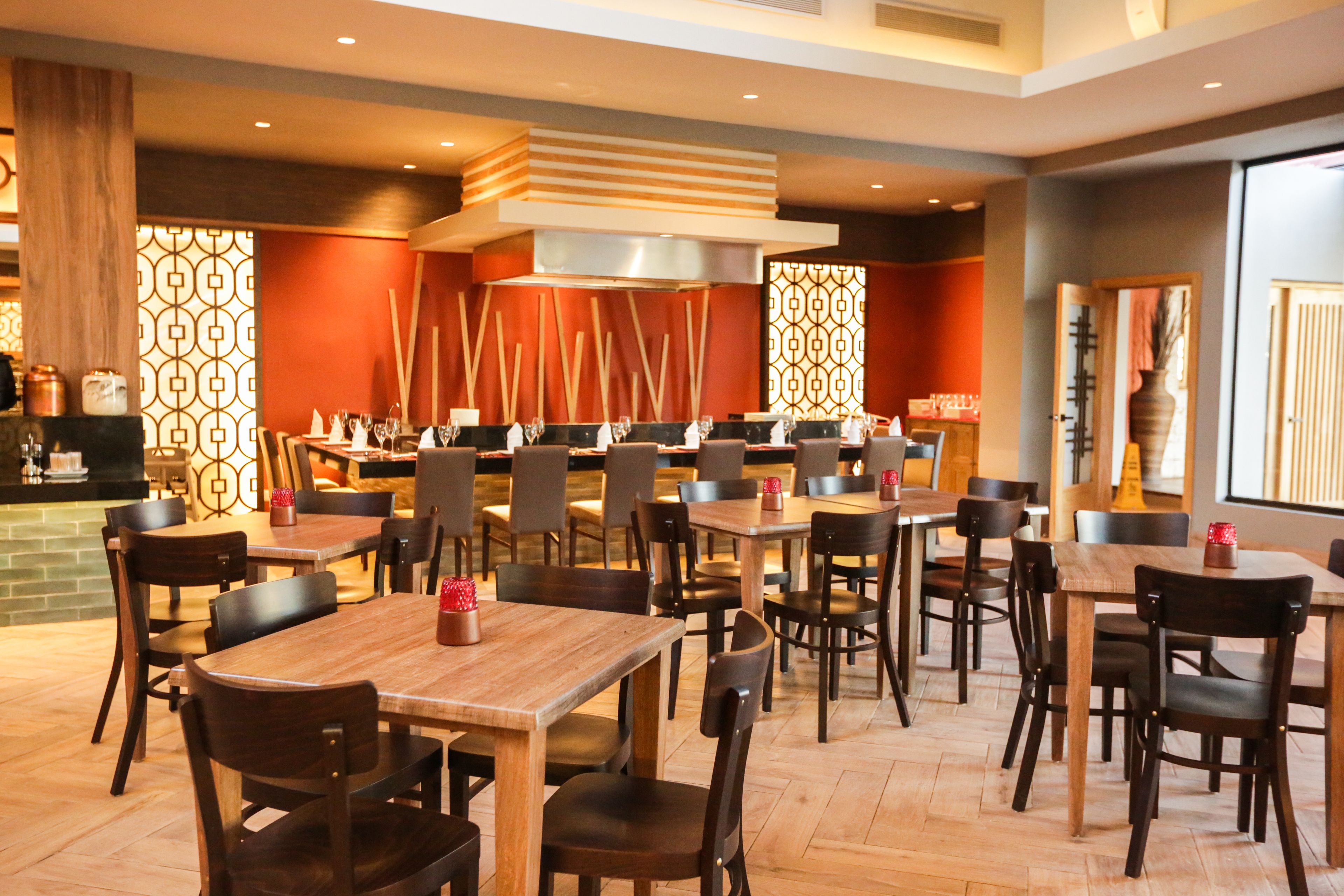 Un restaurante con varias mesas y sillas de madera, una zona de cocina central y paneles decorativos en las paredes.