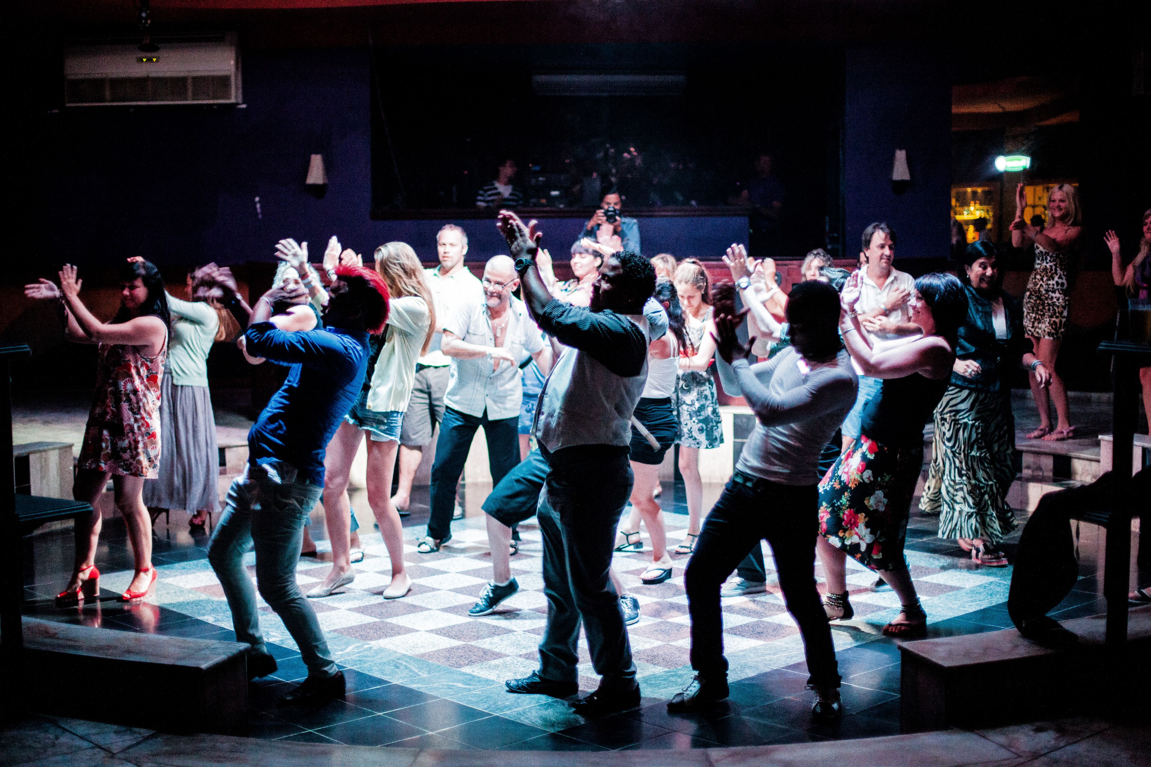 A group of people dancing on a checkered floor in a dimly lit room.