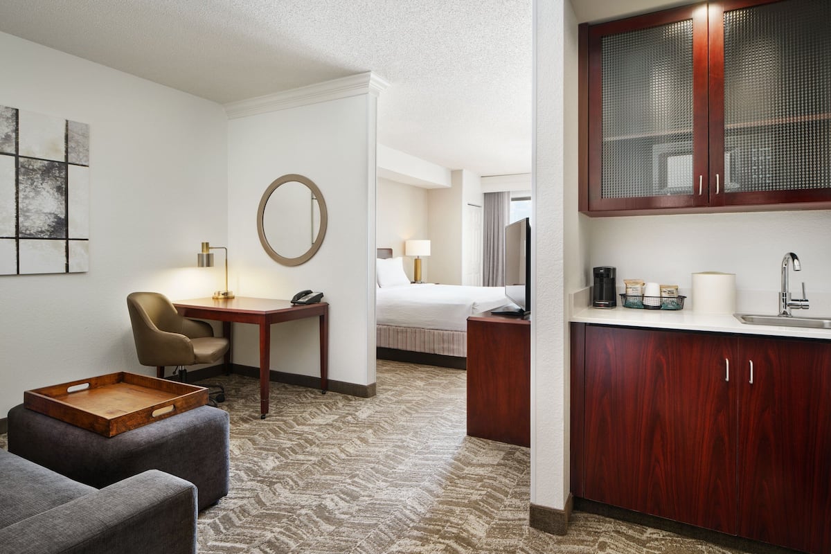 Quarto de hotel com cama, escrivaninha com luminária, cadeira, mesinha com bandeja e área de cozinha compacta com pia e armários.