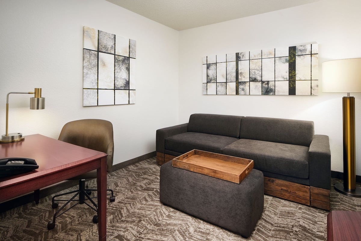Quarto de hotel moderno com sofá cinza, mesa lateral de madeira, escrivaninha com abajur e arte abstrata na parede.