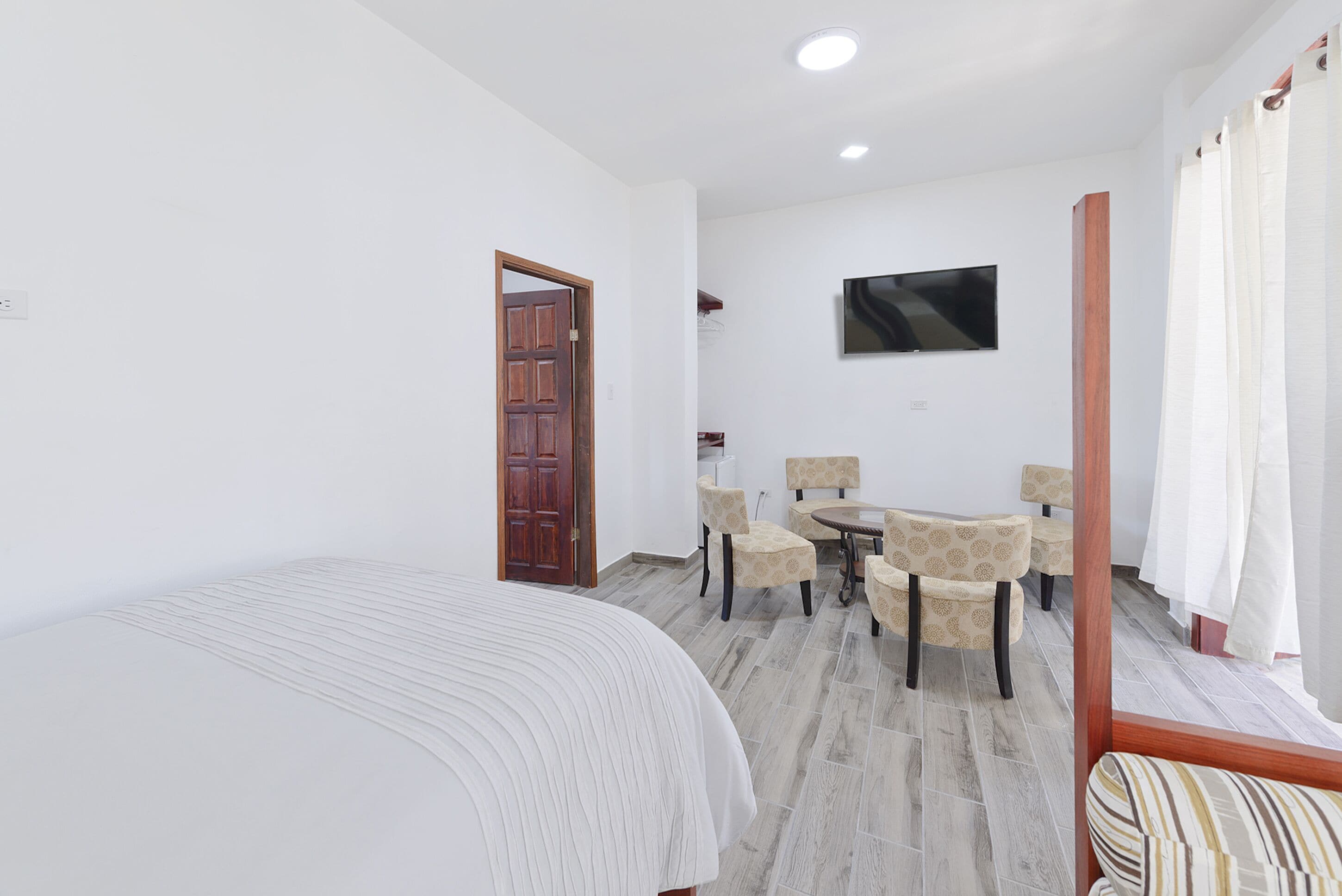 Een slaapkamer met een bed, een houten deur, een eethoek met stoelen, een tafel en een aan de muur bevestigde televisie.