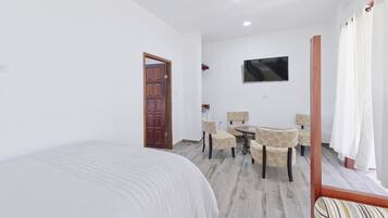 Un dormitorio con cama, puerta de madera, zona de comedor con sillas, una mesa y televisor montado en la pared.