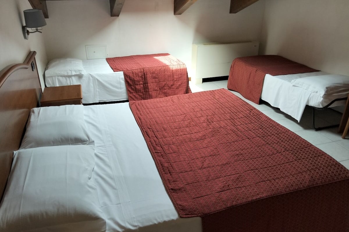 Ein Hotelzimmer mit zwei Betten, jedes mit roter Bettdecke und weißen Laken. Es gibt ein Holz-Kopfstück, einen Nachttisch und eine Wandlampe.