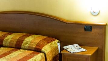 A neatly made bed with a striped quilt, a wooden nightstand with a newspaper, and a wall-mounted light fixture.